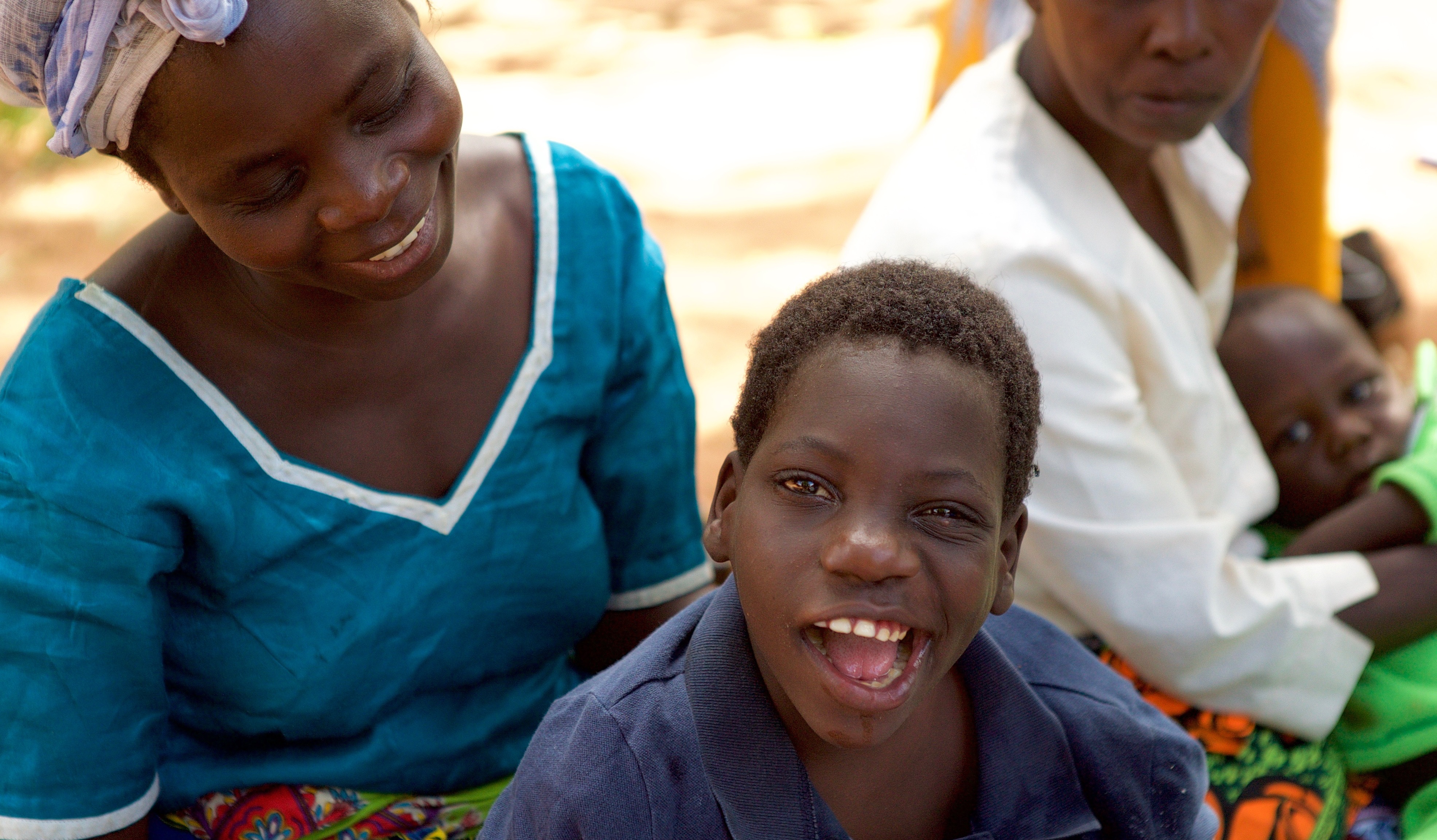 A Zambia woman sits behind her son, looking at him. He looks at the camera, smiling.